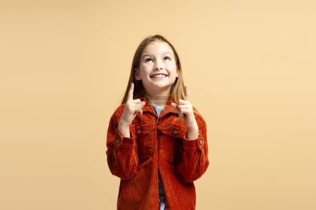 Téléchargez les photos : Jolie petite fille souriante portant des vêtements décontractés élégants pointant les doigts vers le haut, regardant isolé sur fond beige. Concept de publicité - en image libre de droit