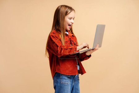 Téléchargez les photos : Portrait de souriante petite fille excitée tenant à l'aide d'un ordinateur portable, tapant sur le clavier, faisant des devoirs debout isolé sur fond beige. Concept technologique - en image libre de droit