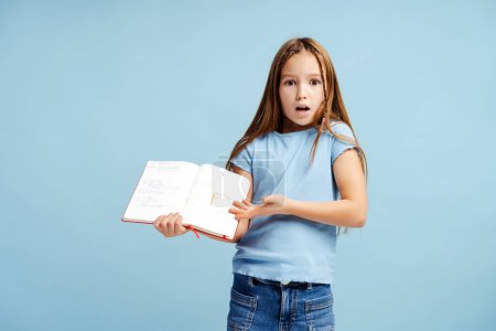 Foto de Retrato de una niña triste y linda sosteniendo un cuaderno con mala nota, de pie aislado sobre fondo azul. Regreso al concepto escolar - Imagen libre de derechos