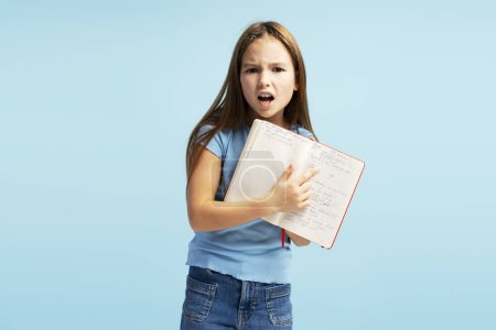 Foto de Niña enojada alumna sosteniendo un cuaderno apuntando con el dedo mirando a la cámara de pie aislada sobre fondo azul. Regreso al concepto escolar - Imagen libre de derechos