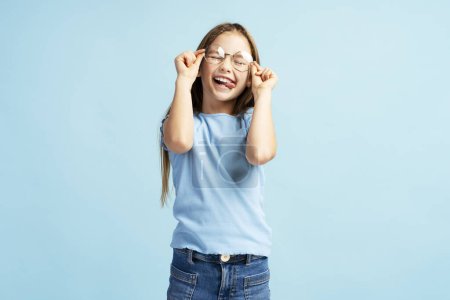 Foto de Sonriente niña linda con gafas con estilo, jugando con los ojos cerrados, de pie aislado sobre fondo azul. Concepto de infancia - Imagen libre de derechos