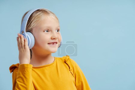 Téléchargez les photos : Portrait de mignonne belle petite fille souriante portant des écouteurs sans fil regardant loin sur l'espace de copie, debout isolé sur fond bleu. Concept publicitaire, technologie - en image libre de droit