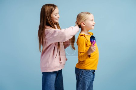 Téléchargez les photos : Jolie belle fille brossant les cheveux de sa petite sœur, faisant sa coiffure, portant des vêtements colorés, debout isolé sur fond bleu. Concept d'amour, de famille, de soins - en image libre de droit