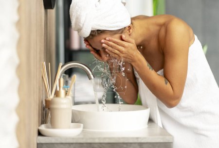 Téléchargez les photos : Belle femme portant une serviette blanche après la douche, le visage de lavage avec de l'eau debout dans la salle de bain élégante à la maison. Concept de routine du matin, soin de la peau - en image libre de droit