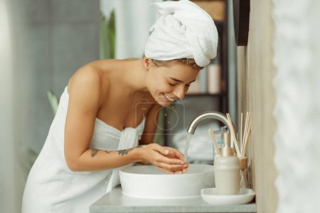 Foto de Retrato de una joven mujer alegre usando toalla blanca después de la ducha, lavando la cara con agua de pie en el elegante baño en casa. Concepto de rutina matinal - Imagen libre de derechos