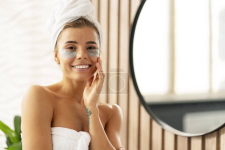 Foto de Mujer atractiva feliz con parches para los ojos después de la ducha, con toalla blanca, de pie en el baño cerca del espejo mirando a la cámara. Concepto de cuidado de la piel - Imagen libre de derechos