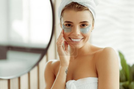 Téléchargez les photos : Belle femme souriante avec des patchs après la douche, portant une serviette blanche, debout dans la salle de bain près du miroir regardant la caméra. Soins de la peau, concept de beauté naturelle - en image libre de droit