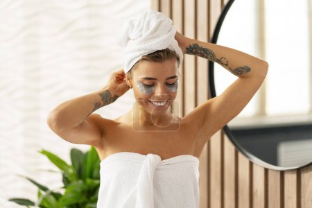 Téléchargez les photos : Femme positive gaie avec des patchs oculaires après la douche, portant une serviette blanche, debout dans la salle de bain près du miroir. Concept de routine du matin, soin de la peau - en image libre de droit