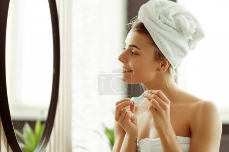 Téléchargez les photos : Souriant femme heureuse avec serviette sur la tête appliquant des patchs, regardant dans le miroir debout dans la salle de bain élégante à la maison. Concept spa, soin visage anti-âge - en image libre de droit