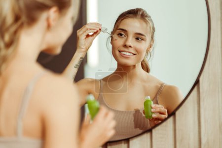 Téléchargez les photos : Belle jeune femme tenant bouteille verte appliquant du sérum sur le visage, regardant dans le miroir, debout dans la salle de bain à la maison. Concept de soin de la peau, procédure cosmétique, hydratation - en image libre de droit
