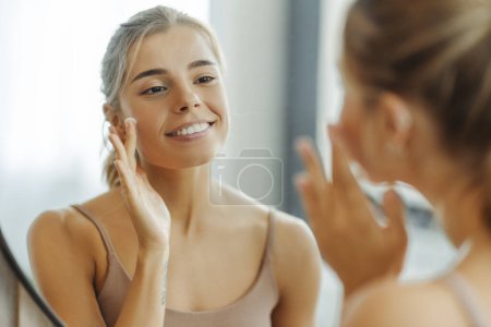 Photo for Beautiful young woman applying cream on face, standing in bathroom at home. Concept of skin care, moisturizing, hydration - Royalty Free Image