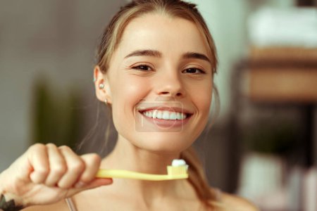 Téléchargez les photos : Souriant jeune femme gaie tenant une brosse à dents avec du dentifrice, regardant la caméra, brossant les dents debout dans la salle de bain à la maison. Concept de soins dentaires - en image libre de droit