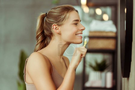 Foto de Mujer joven sonriente con camiseta sin mangas sosteniendo el cepillo de dientes con pasta de dientes, mirándose en el espejo, cepillándose los dientes en el baño en casa. Concepto de cuidado dental, tratamiento - Imagen libre de derechos