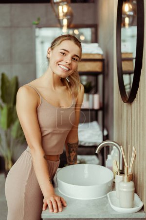 Photo for Positive beautiful young woman wearing tank top and pants, looking at camera, standing in bathroom near mirror at home. Natural beauty, morning routine, body care - Royalty Free Image