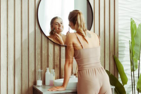 Téléchargez les photos : Belle jeune femme confiante portant débardeur et pantalon faisant un massage à rouleaux, regardant dans le miroir, debout dans une élégante salle de bain moderne à la maison. Concept de soin de la peau, beauté naturelle - en image libre de droit