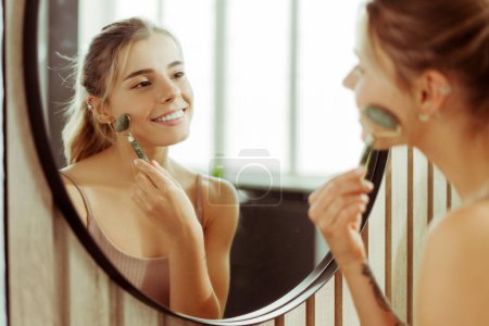 Téléchargez les photos : Portrait de jeune femme positive en débardeur, regardant dans le miroir, faisant un massage à rouleaux, levant, debout dans la salle de bain. Concept de soin de la peau, routine matinale - en image libre de droit