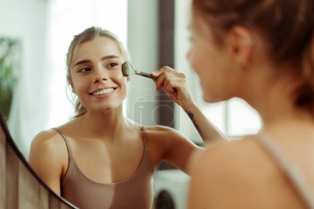 Téléchargez les photos : Portrait de belle jeune femme en débardeur, regardant dans le miroir, faisant un massage à rouleaux, levant, debout dans la salle de bain. Concept de soin de la peau, procédure cosmétique, routine matinale - en image libre de droit