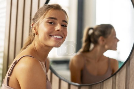 Téléchargez les photos : Portrait de femme confiante et positive portant un débardeur, avec un maquillage parfait regardant la caméra debout dans la salle de bain à la maison. Concept de soins de la peau - en image libre de droit