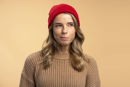 Foto de Retrato de una mujer joven y seria, hipster con un elegante sombrero rojo, suéter marrón mirando hacia otro lado aislado sobre fondo beige. Concepto de publicidad - Imagen libre de derechos