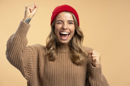 Foto de Retrato de una atractiva mujer sonriente con sombrero rojo y suéter marrón de invierno sosteniendo las manos, gesto de victoria, mirando a la cámara. Concepto de ganar - Imagen libre de derechos