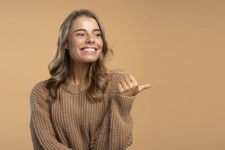 Téléchargez les photos : Portrait d'une belle femme souriante portant un pull marron pointant du doigt l'espace de copie, regardant ailleurs. Concept de publicité - en image libre de droit