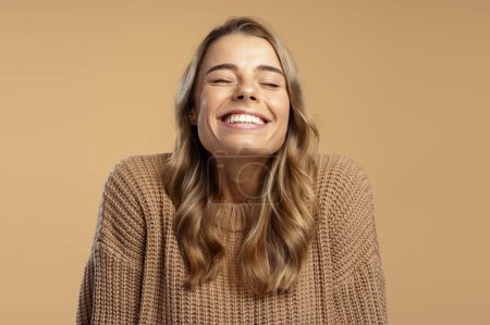Foto de Retrato de mujer joven positiva con los ojos cerrados sonriendo, vistiendo suéter marrón acogedor aislado sobre fondo beige, primer plano. Concepto dental - Imagen libre de derechos