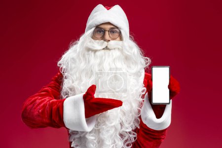 Téléchargez les photos : Funny Santa Claus tenant téléphone mobile montrant écran blanc regardant la caméra isolée sur fond rouge, maquette. Achats en ligne, vente, concept de Noël - en image libre de droit