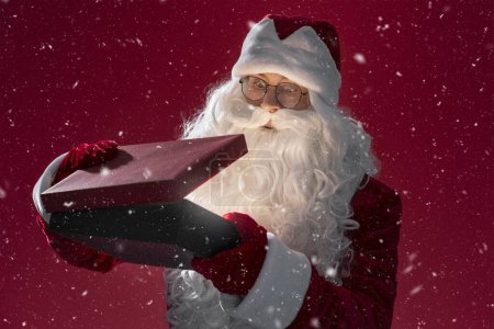 Téléchargez les photos : Funny Santa Claus tenant et ouvrant boîte cadeau isolé sur fond rouge. Vacances, Nouvel An, concept magique - en image libre de droit