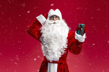 Téléchargez les photos : Homme drôle portant costume du Père Noël tenant boîte cadeau en regardant la caméra isolée sur fond rouge. Joyeux Noël, concept de vente d'hiver - en image libre de droit