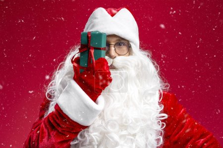 Photo for Santa Claus holding gift box near face isolated on red background. Holiday, Merry Christmas concept - Royalty Free Image