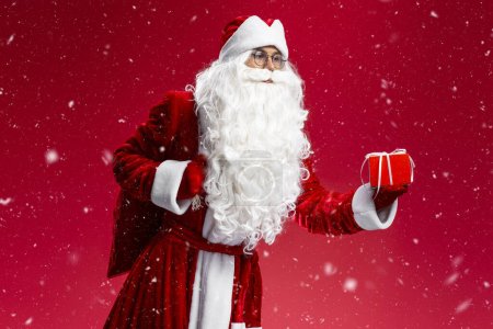 Téléchargez les photos : Père Noël tenant le sac, transporter boîte cadeau isolé sur fond rouge. Vacances, Noël, concept publicitaire - en image libre de droit