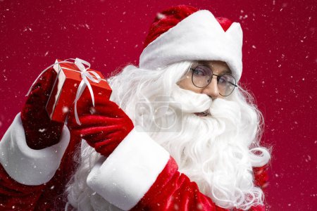 Foto de Primer plano retrato de Santa Claus sosteniendo caja de regalo mirando a la cámara aislada sobre fondo rojo. Feliz Año Nuevo, concepto de Navidad - Imagen libre de derechos