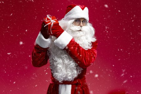Photo for Santa Claus holding gift box looking at camera isolated on red background. Happy New Year, Christmas, holiday, winter concept - Royalty Free Image