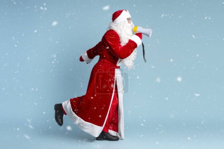 Photo for Portrait of funny Santa holding megaphone running, hurry up announcing winter sale isolated on blue background, copy space. Christmas, holidays, advertisement concept - Royalty Free Image