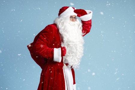 Foto de Santa Claus sostiene la bolsa con regalos mirando hacia otro lado, se apresura aislado sobre fondo azul con nieve, espacio para copiar. Vacaciones, concepto de publicidad - Imagen libre de derechos
