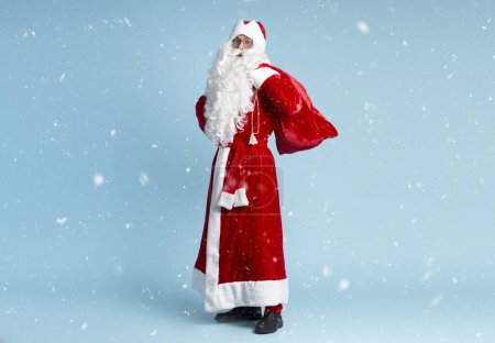 Photo for Saint Nicholas holding red bag with gifts looking at camera isolated on blue background with snow. Winter holidays concept - Royalty Free Image