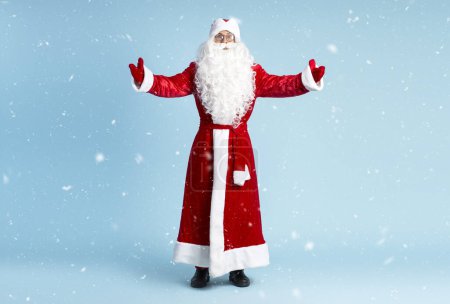 Foto de Santa Claus sosteniendo las manos abiertas, abrazándose mirando a la cámara aislada sobre fondo azul con nieve. Concepto de vacaciones de invierno - Imagen libre de derechos