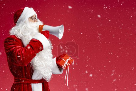 Foto de Retrato de Santa Claus sosteniendo caja de regalo y megáfono anunciando las ventas de invierno aisladas sobre fondo rojo, espacio de copia. Vacaciones, Navidad, concepto de publicidad - Imagen libre de derechos