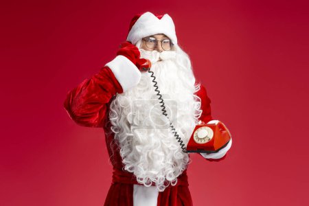 Photo for Santa Claus holding retro telephone answering call isolated on red background - Royalty Free Image