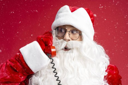 Téléchargez les photos : Gros plan Père Noël tenant rétro téléphone répondre à l'appel, souhait d'écoute isolé sur fond rouge. Concept vacances d'hiver - en image libre de droit