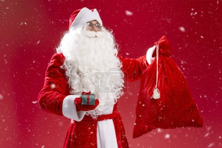 Foto de Retrato de Santa Claus bolsa de sujeción, llevar caja de regalo aislado sobre fondo rojo. Vacaciones, celebración de Navidad, concepto de publicidad - Imagen libre de derechos