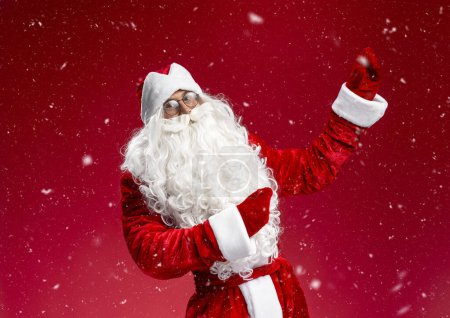Foto de Feliz Santa Claus bailando, divirtiéndose apuntando con las manos a la nieve aislada sobre fondo azul. Navidad, concepto de vacaciones de invierno - Imagen libre de derechos