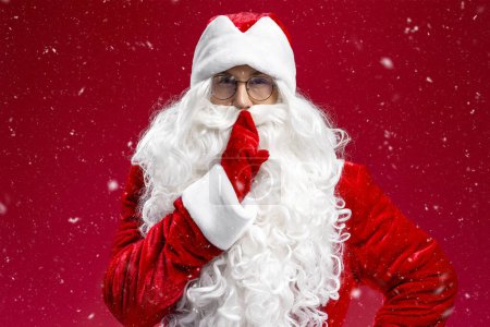 Photo for Portrait of Santa Claus holding finger on lips looking at camera isolated on red background - Royalty Free Image