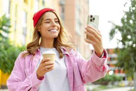 Téléchargez les photos : Jolie femme souriante portant un chapeau élégant rouge tenant une tasse de café et un smartphone prenant un selfie dans la rue. Influenceur utilisant la vidéo d'enregistrement d'application mobile, communication en ligne dans le parc - en image libre de droit