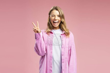 Foto de Beautiful smiling blonde woman with wavy silky hair wearing stylish pink shit winks, showing victory sign isolated on background. Portrait of happy successful influencer greeting looking at camera - Imagen libre de derechos
