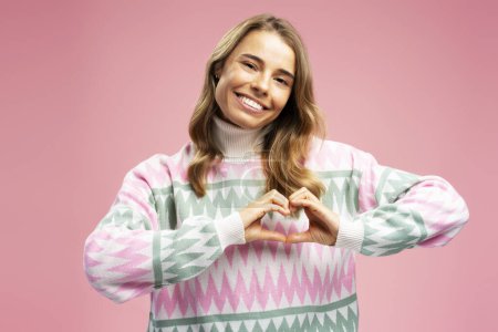 Foto de Mujer hermosa sonriente, linda influencer alegre usando suéter de invierno elegante que muestra la forma del corazón para los suscriptores aislados en el fondo rosa. Estilo de vida positivo, concepto de redes sociales - Imagen libre de derechos