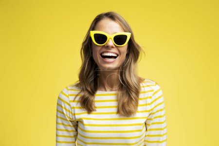Foto de Mujer joven sonriente con gafas de sol elegantes mirando a la cámara aislada sobre fondo amarillo. Modelo de moda posando para fotos en el estudio. Verano, viajes, concepto de vacaciones - Imagen libre de derechos