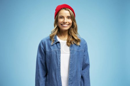Téléchargez les photos : Portrait de belle femme souriante portant un chapeau hipster rouge et une veste en jean élégante isolée sur fond bleu. Touriste féminine aux cheveux ondulés et soyeux regardant la caméra. Concept de voyage - en image libre de droit