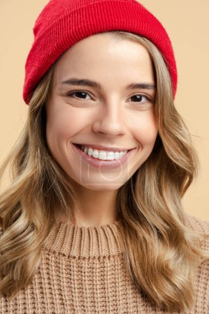Foto de Primer plano retrato sonriente mujer atractiva con sombrero hipster rojo y suéter de invierno con estilo mirando a la cámara aislada sobre fondo beige. Estilo de vida positivo, concepto de belleza natural - Imagen libre de derechos