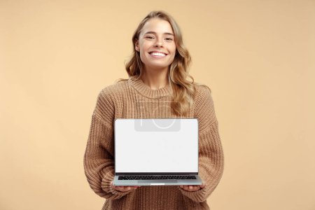 Téléchargez les photos : Femme souriante tenant un ordinateur portable montrant écran d'ordinateur isolé sur fond beige, maquette. Étudiant heureux intelligent étudiant, apprendre la langue, concept d'éducation en ligne. Technologie, site web - en image libre de droit